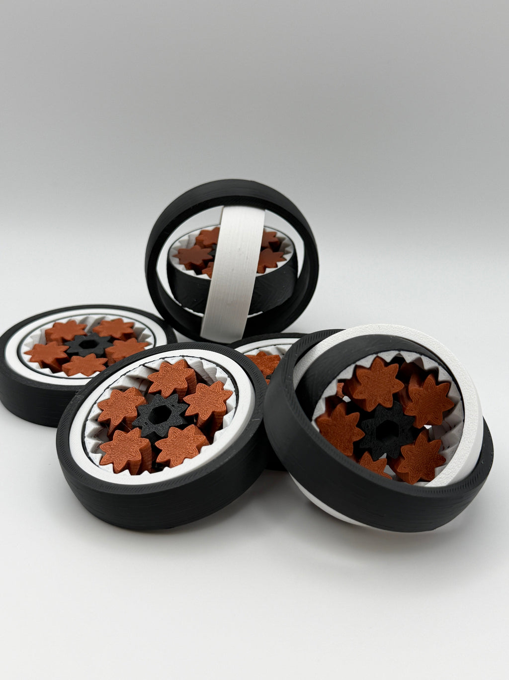 Five gyro spinning fidget toys. A black exterior layer, followed by a white layer, followed by another black layer that spins. 5 bronze coloured star shape wheels wrapped around a black star shape cog that spins the bronze wheels. 