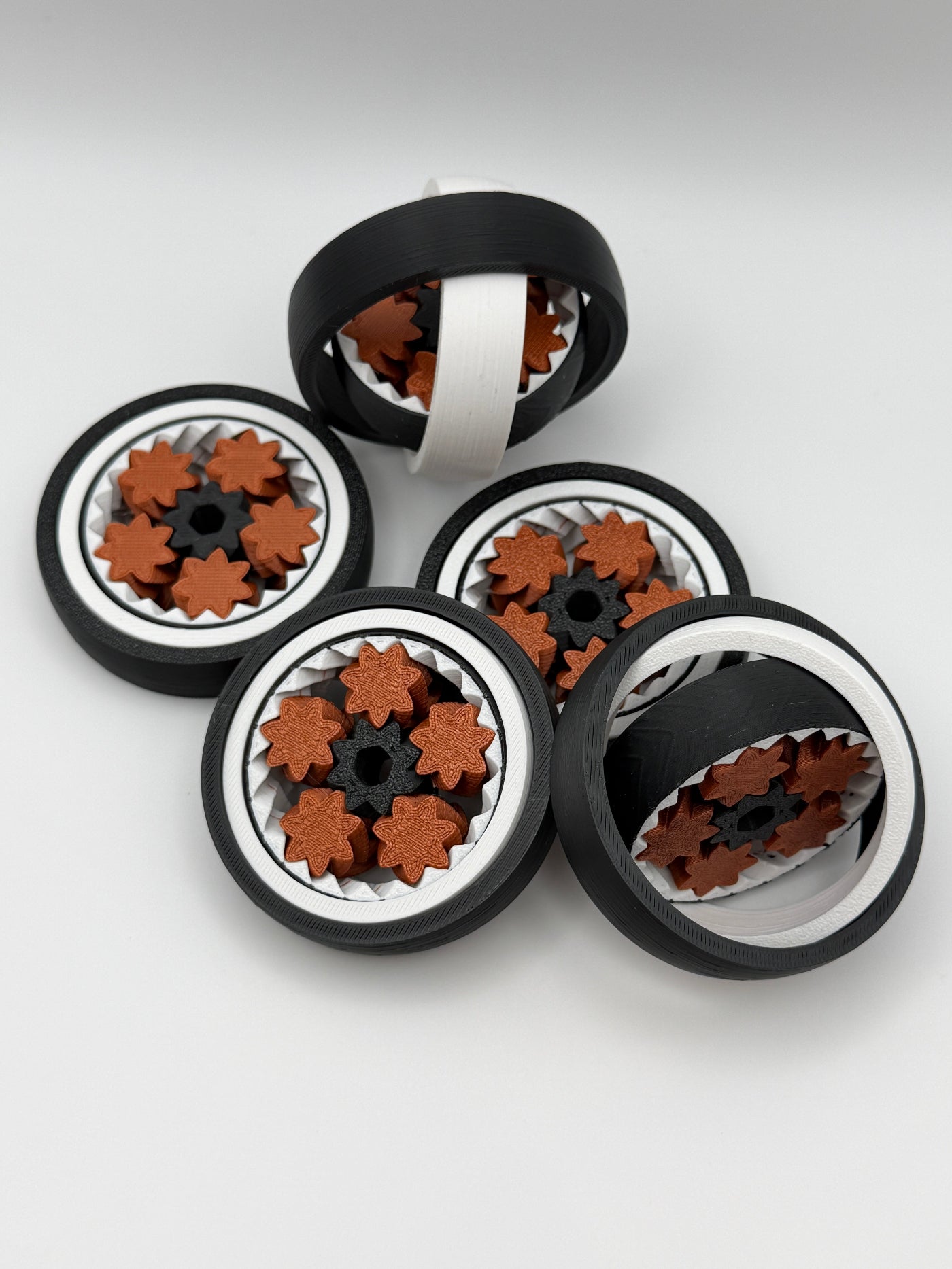 Five gyro spinning fidget toys. A black exterior layer, followed by a white layer, followed by another black layer that spins. 5 bronze coloured star shape wheels wrapped around a black star shape cog that spins the bronze wheels. 