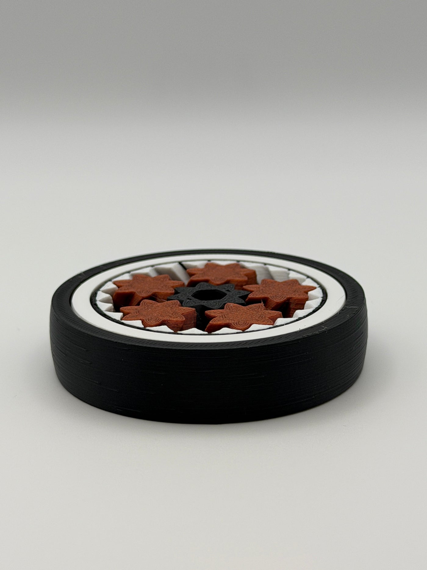 A gyro spinning fidget toy. With a black exterior layer, followed by a white layer, followed by another black layer that spins. 5 bronze coloured star shape wheels wrapped around a black star shape cog that spins the bronze wheels. 