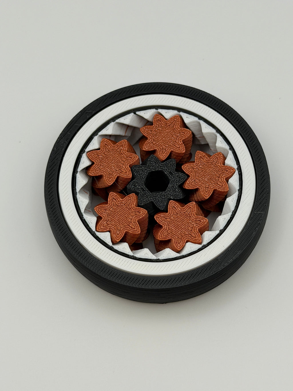 A gyro spinning fidget toy. With a black exterior layer, followed by a white layer, followed by another black layer that spins. 5 bronze coloured star shape wheels wrapped around a black star shape cog that spins the bronze wheels. 