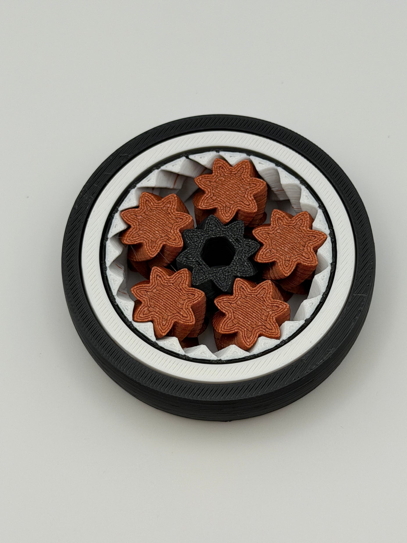 A gyro spinning fidget toy. With a black exterior layer, followed by a white layer, followed by another black layer that spins. 5 bronze coloured star shape wheels wrapped around a black star shape cog that spins the bronze wheels. 