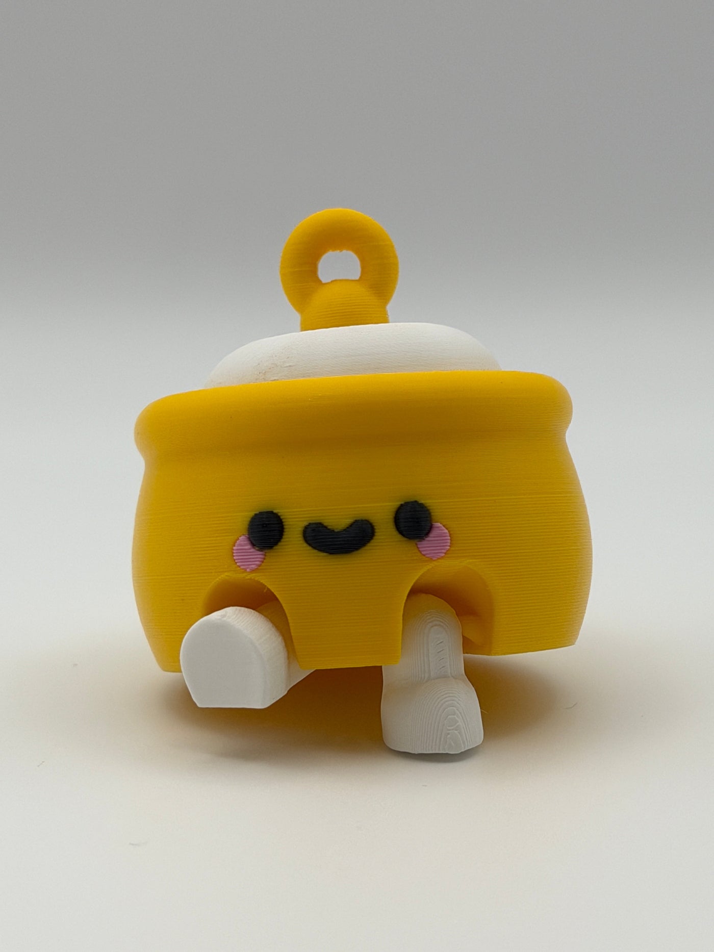 A yellow honey pot with a white fidget clicker top and a yellow keychain attachment with white articulated legs and a black smiley face. 