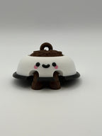 A white dog bowl with brown food and a keychain attachment. The white bowl has a black smiley face and the legs are brown and articulated. 