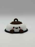 A white dog bowl with brown food and a keychain attachment. The white bowl has a black smiley face and the legs are brown and articulated. 